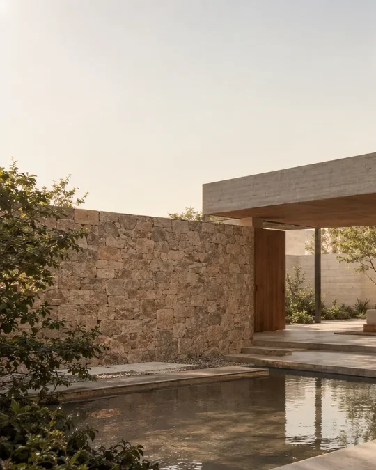 A natural stone wall beside a still reflecting pool, with the edge of a slim timber-and-concrete pavilion catching warm evening light.